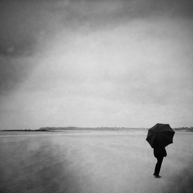 The umbrella, Saint-Malo, Brittany. That day, the wind was blowing hard on the beach and moving sand cloud. The sky was lowering with rain. This young woman suddenly opened her umbrella to brave the elements. An umbrella on the beach in Brittany, this is anecdotal. A funny and graceful time.